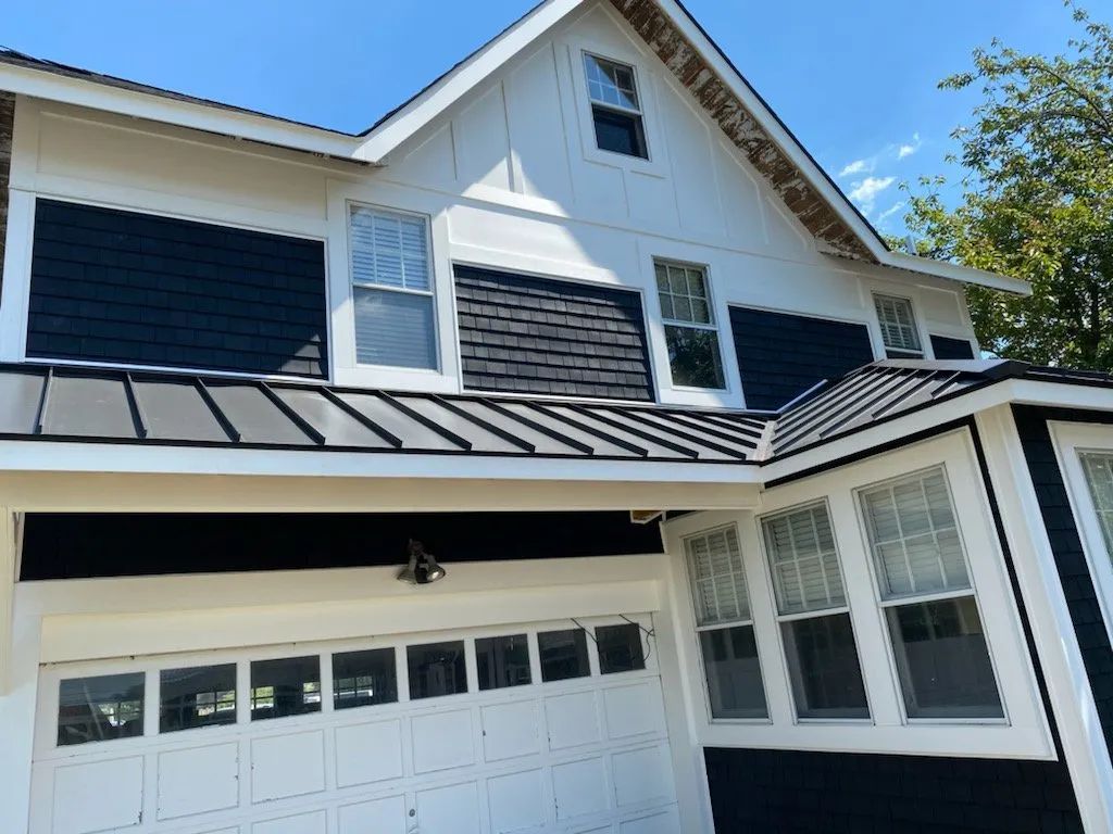 White two-story house exterior with black trim, garage door, and dormer windows under a blue sky