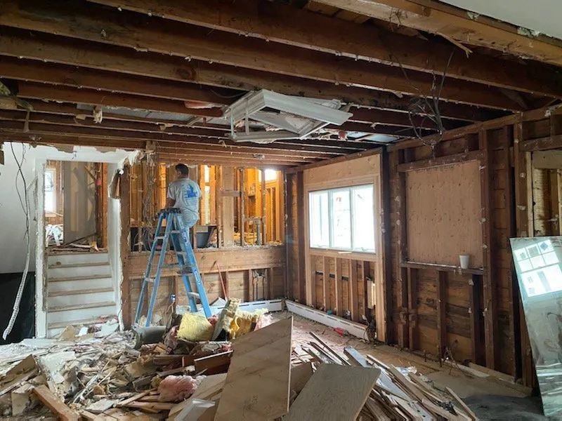 Interior demolition with exposed framing, debris on the floor, and a worker on a ladder.