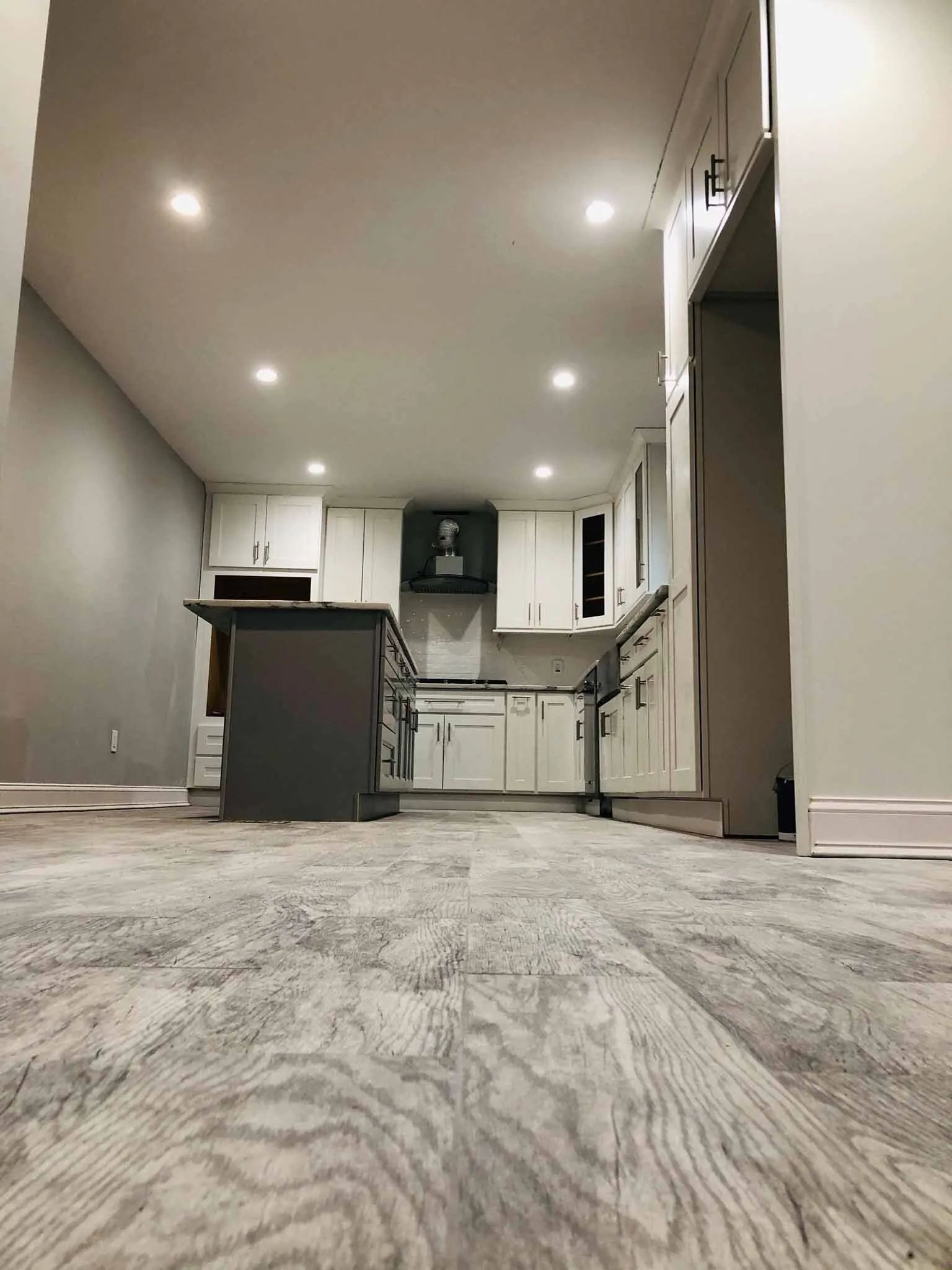 Empty modern kitchen with gray walls, white cabinets, recessed lights, and wood-patterned flooring.