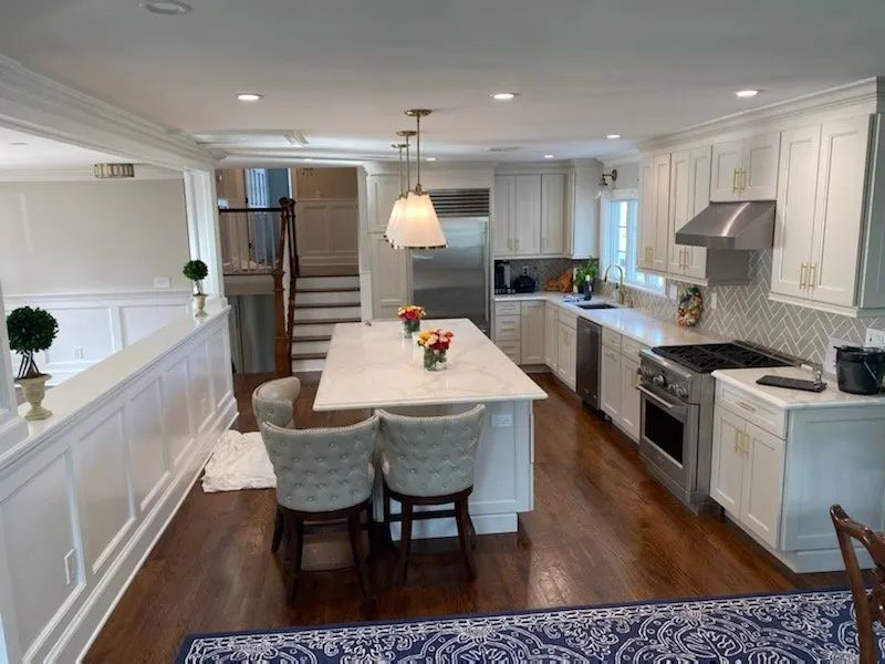 Bright white kitchen with island, gray chairs, stainless appliances, and wood floors leading to stairs.