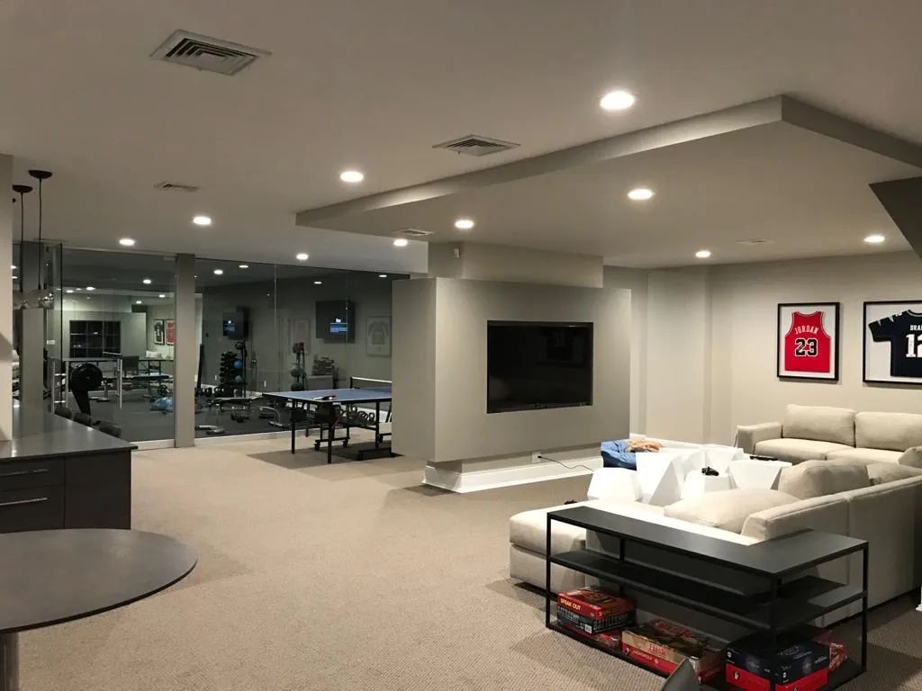 Modern living room with beige sectional, wall-mounted TV, glass coffee table, and framed sports jerseys.