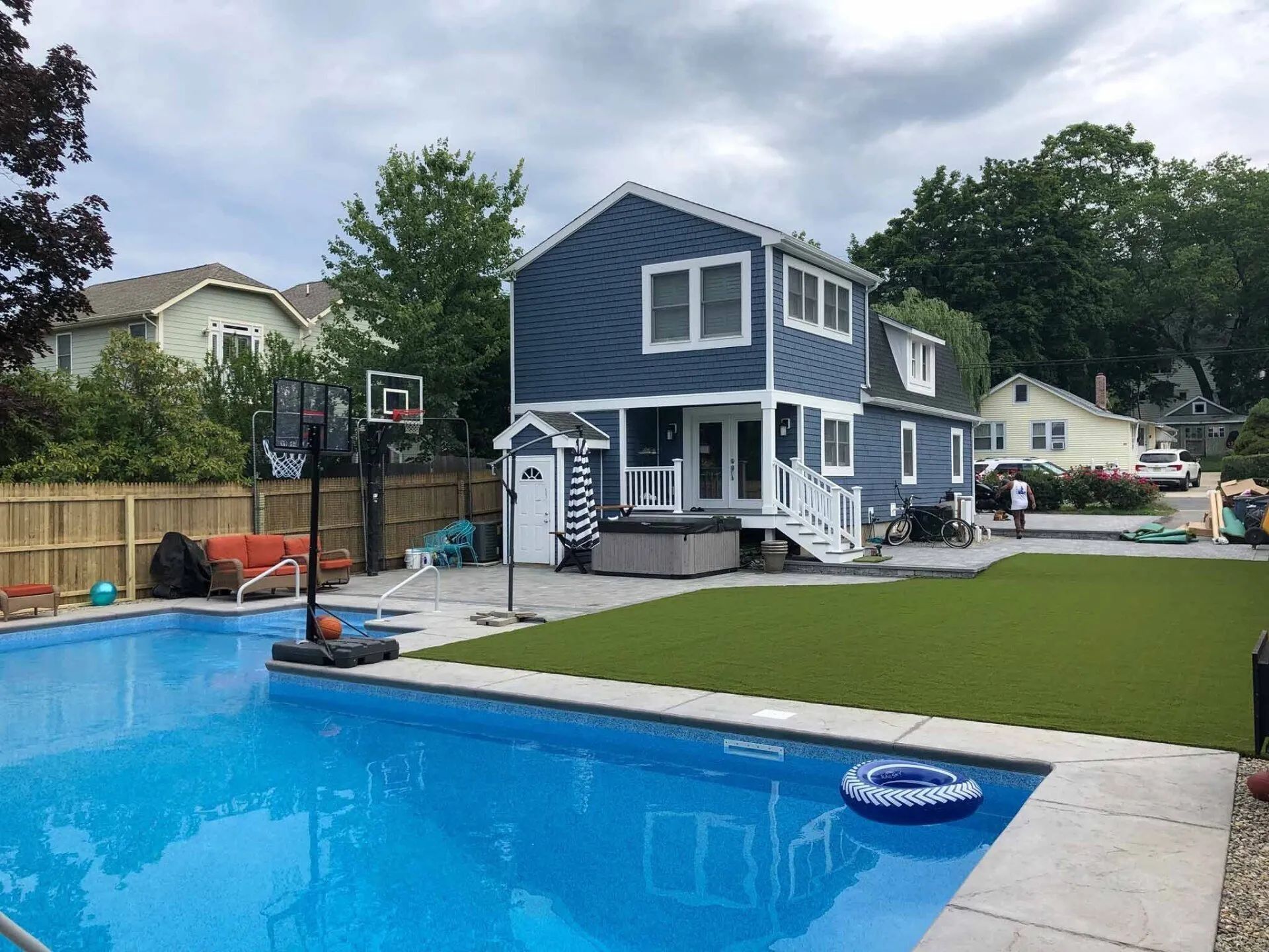 Backyard pool beside a blue two-story house with patio and lawn under cloudy sky