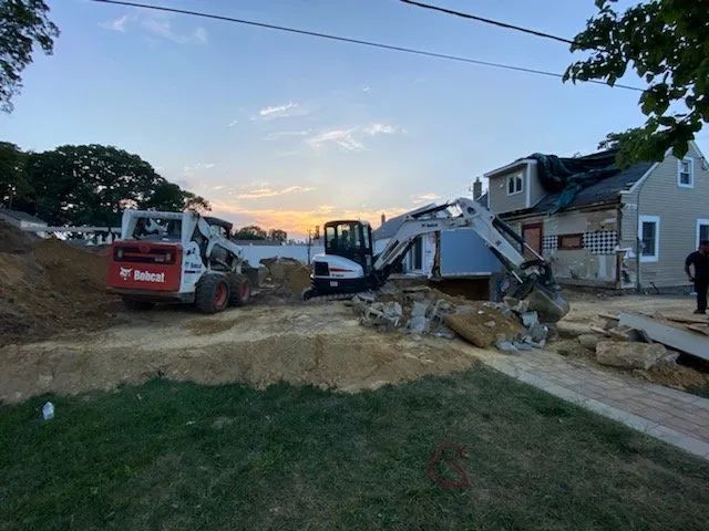 Excavators and a dump truck work on a dirt-filled residential lot at sunset, with houses nearby.