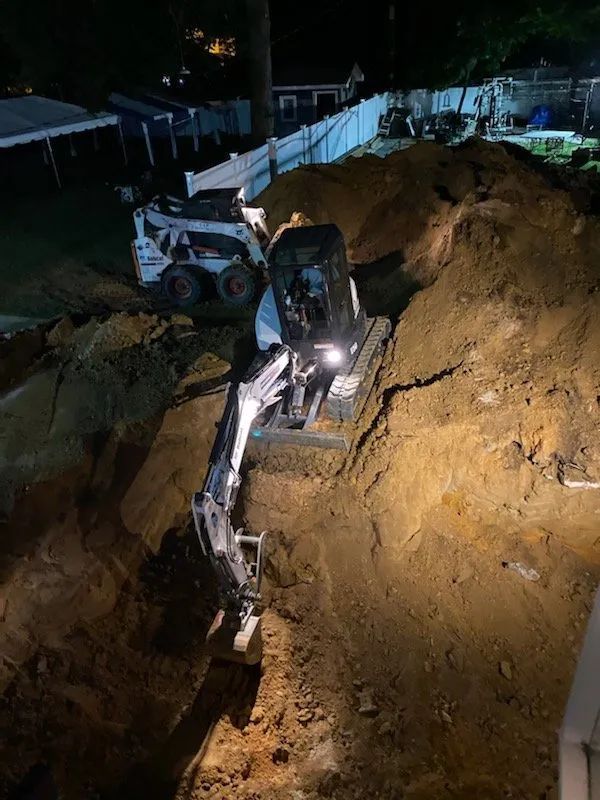 Excavator working in a deep trench at night, with another construction machine nearby and dirt piled along the sides.