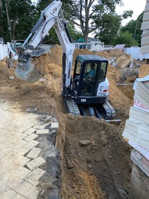 Excavator digging a deep trench at a construction site beside stacked materials and paving stones