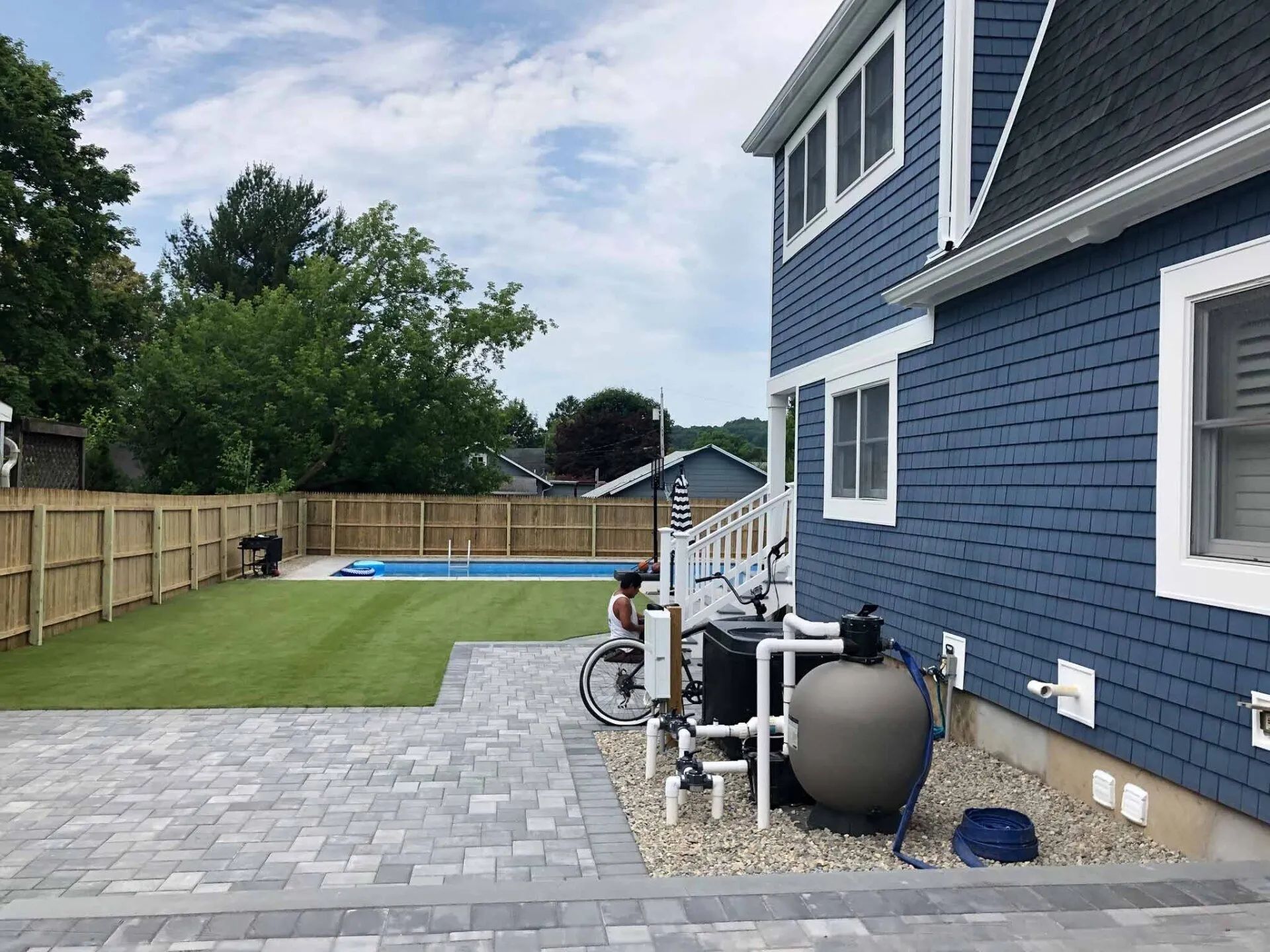 Backyard patio with blue house, stone pavers, green lawn, and a swimming pool along a wooden fence.