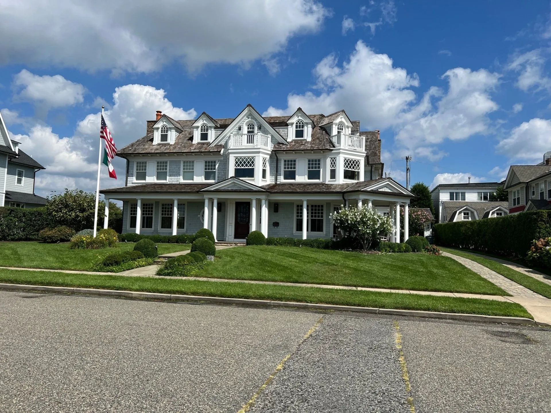 Large white Victorian house with a porch and American flag under a partly cloudy blue sky