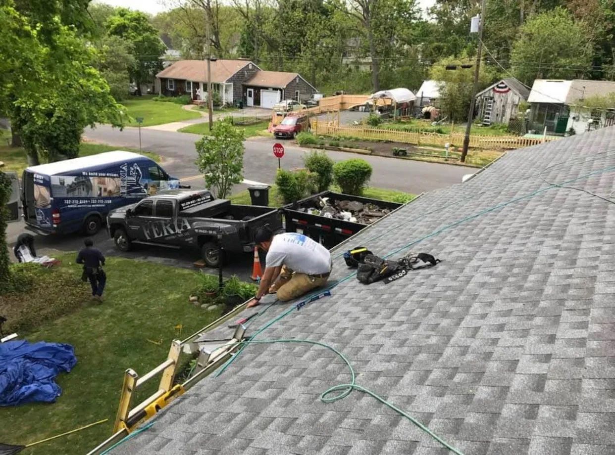 Worker on a roof above a street, with trucks and debris below in a suburban neighborhood.