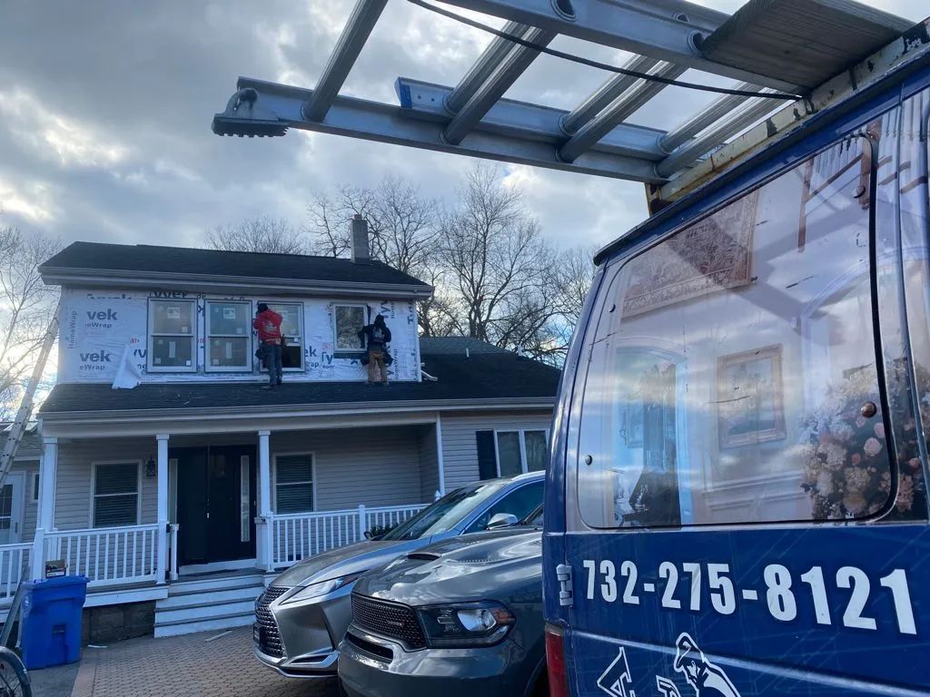 Two-story house with workers on roof and parked car beside a truck canopy with phone number 732-275-8121