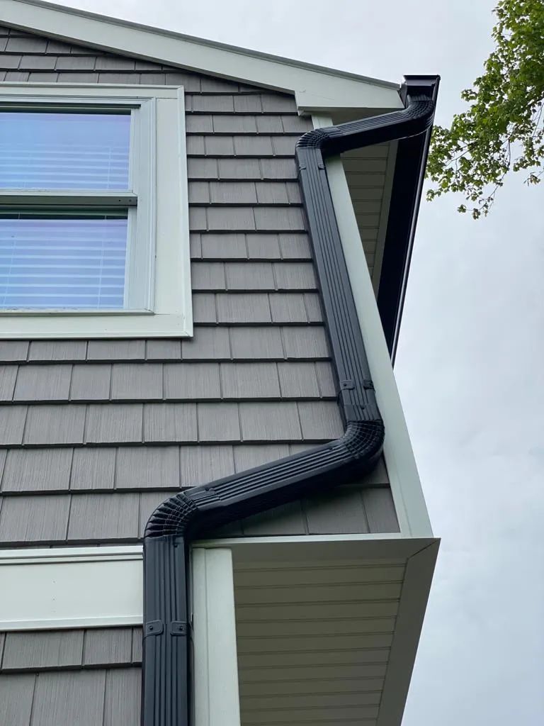 Gray house exterior with a black gutter running down the wall beside a window and roof overhang