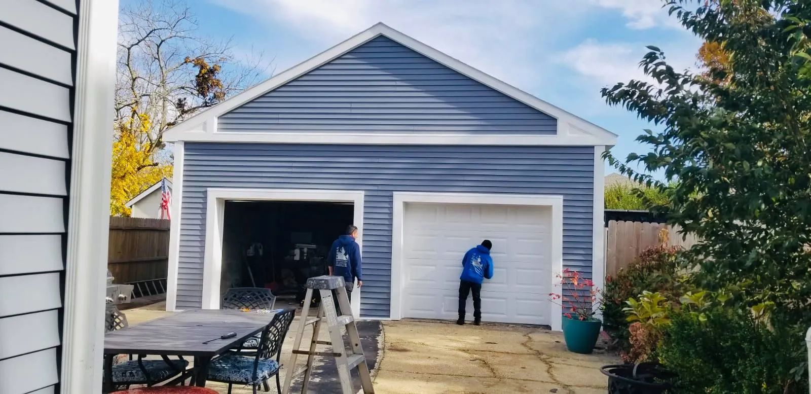 Two workers painting a white two-car garage with open doors in a backyard