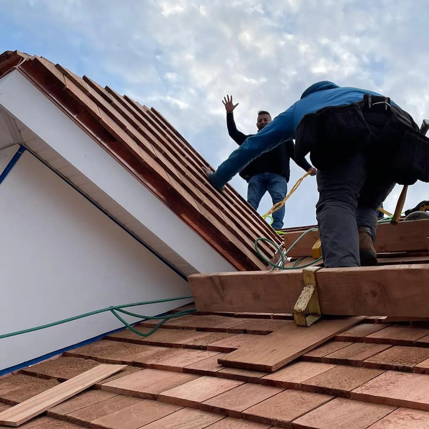 Workers on a roof installing red shingles under a cloudy sky