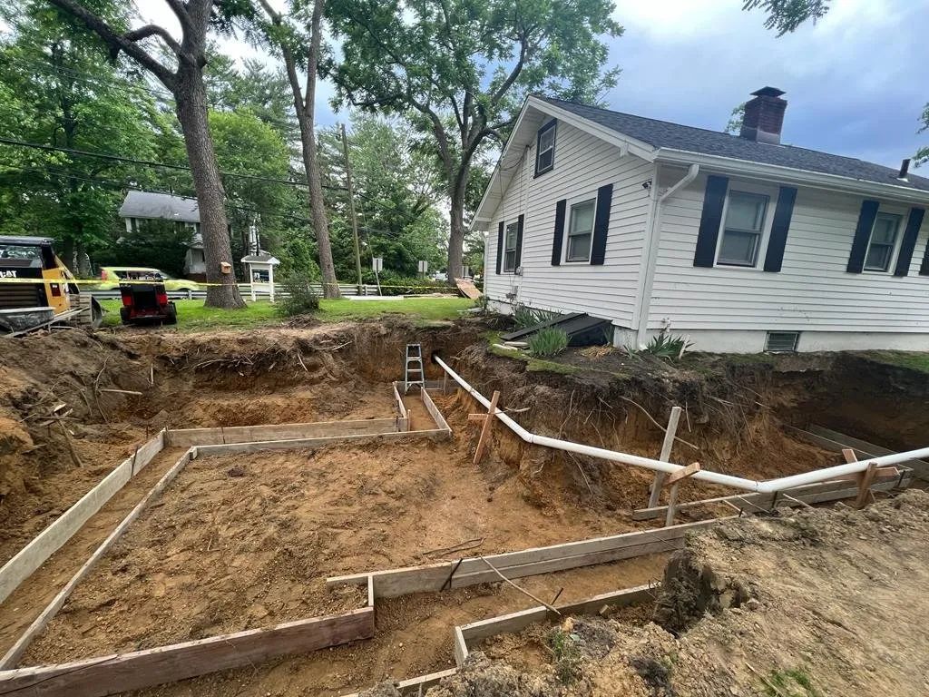 Excavated yard with retaining wall forms beside a white house surrounded by trees.