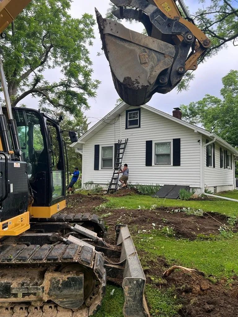 Excavator demolishing a small white house in a yard, with tracks and raised bucket in the foreground.