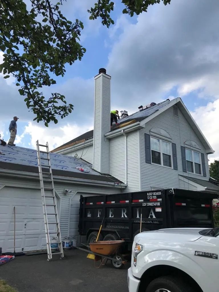 Worker on a roof beside a ladder, chimney, and parked truck at a house under repair.