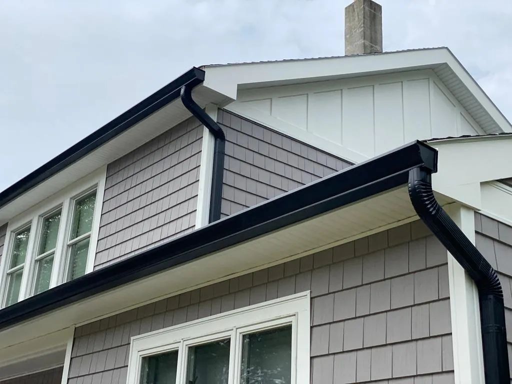 Close-up of a house exterior with gray siding, white trim, and black gutters against a white roofline.