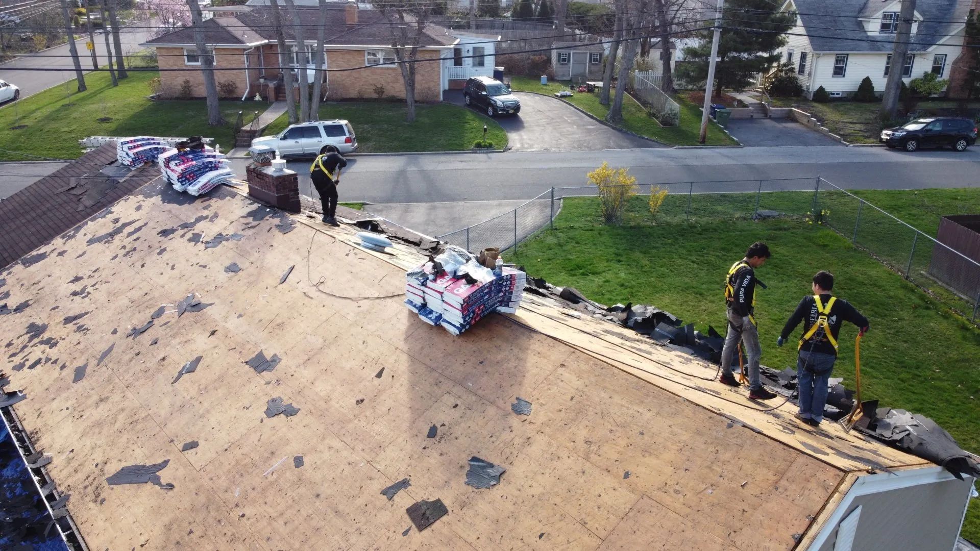 Workers on a shingled roof repairing damage, with a street and houses in the background.