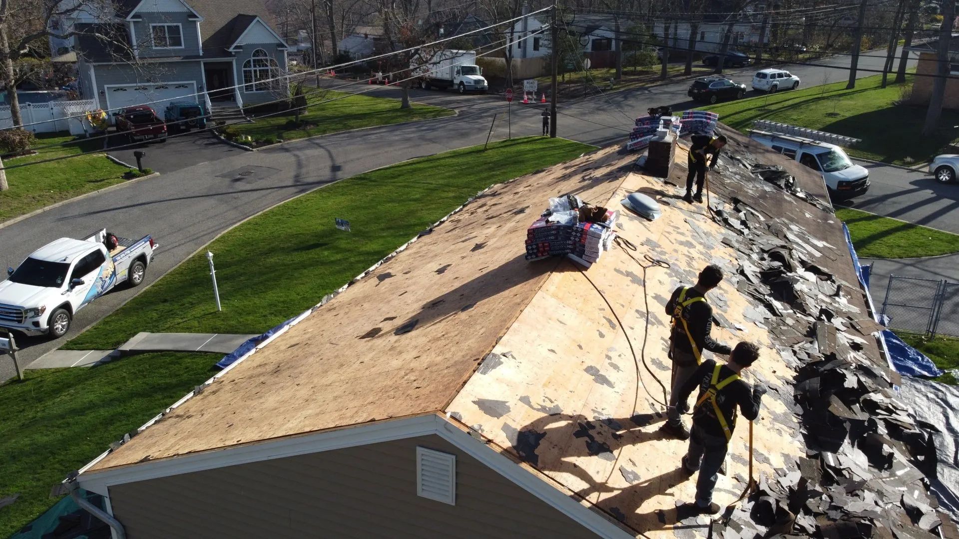 Workers repair a roof with exposed plywood and torn shingles in a suburban neighborhood.