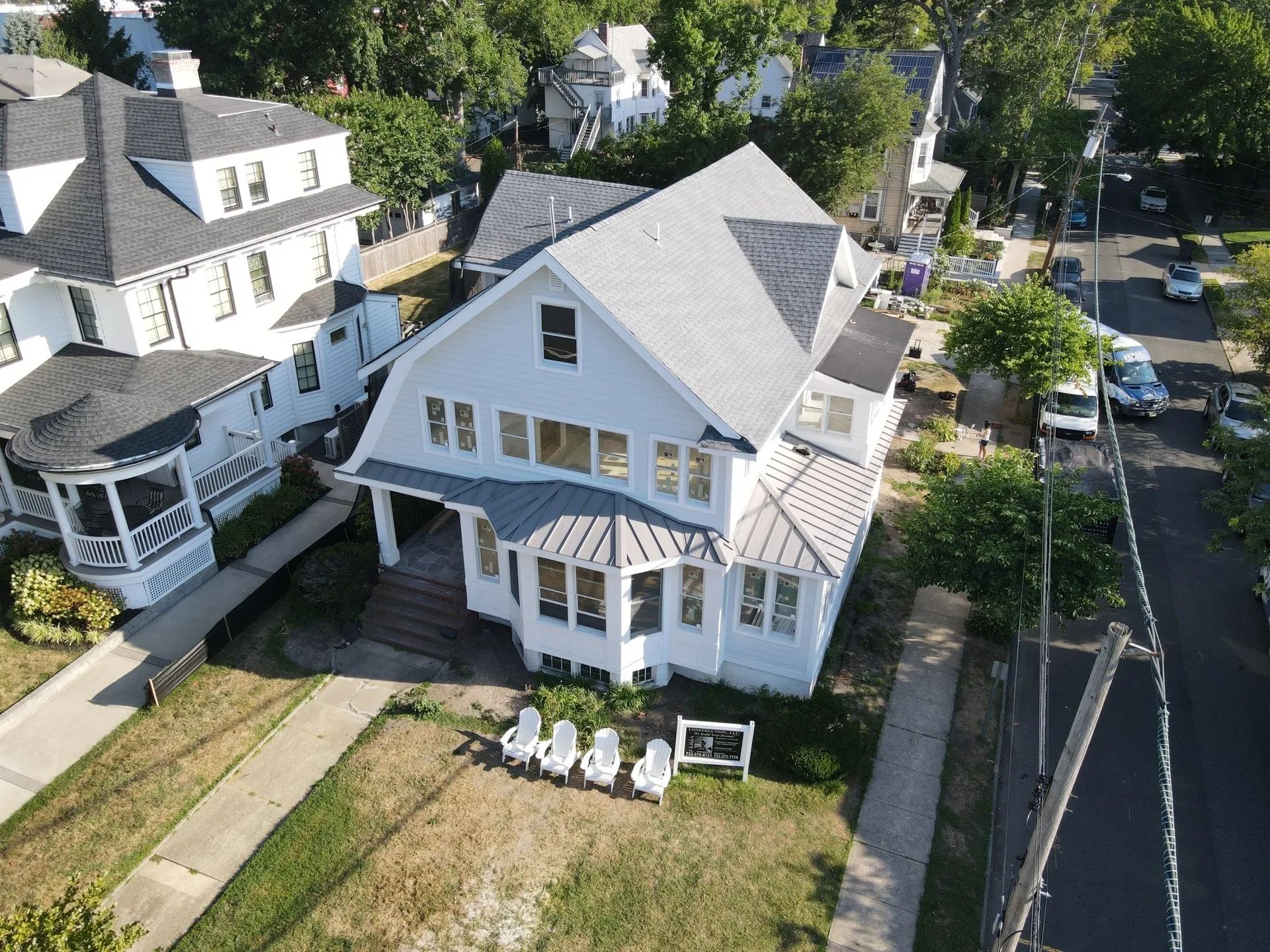 Aerial view of a white house with a front porch and lawn beside a tree-lined residential street.
