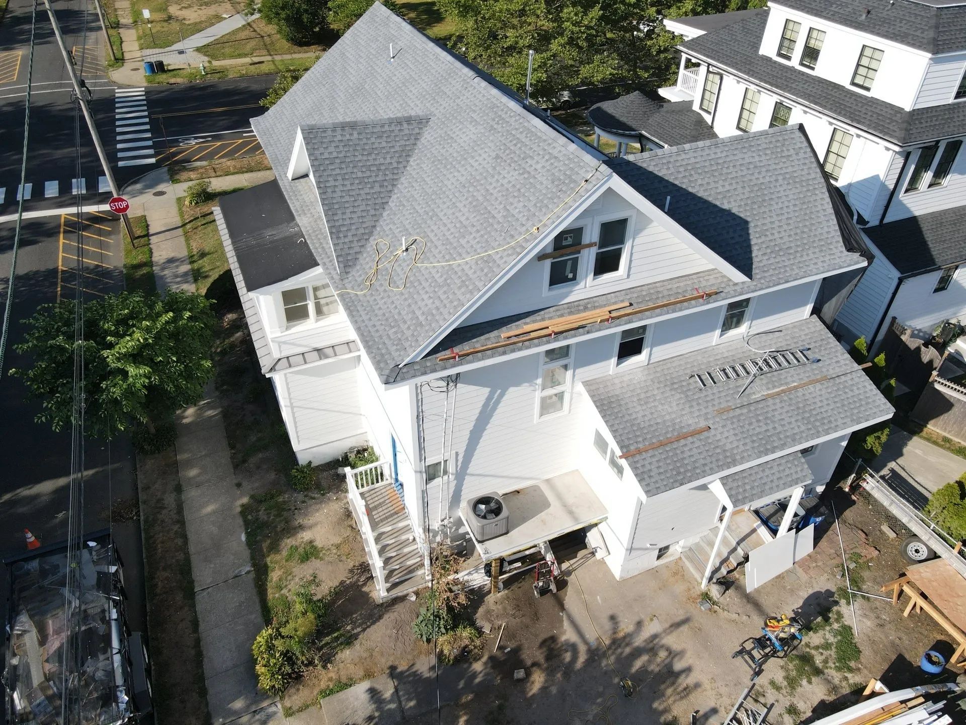 Aerial view of a white house with gray roof, front porch, and yard next to a street and trees.