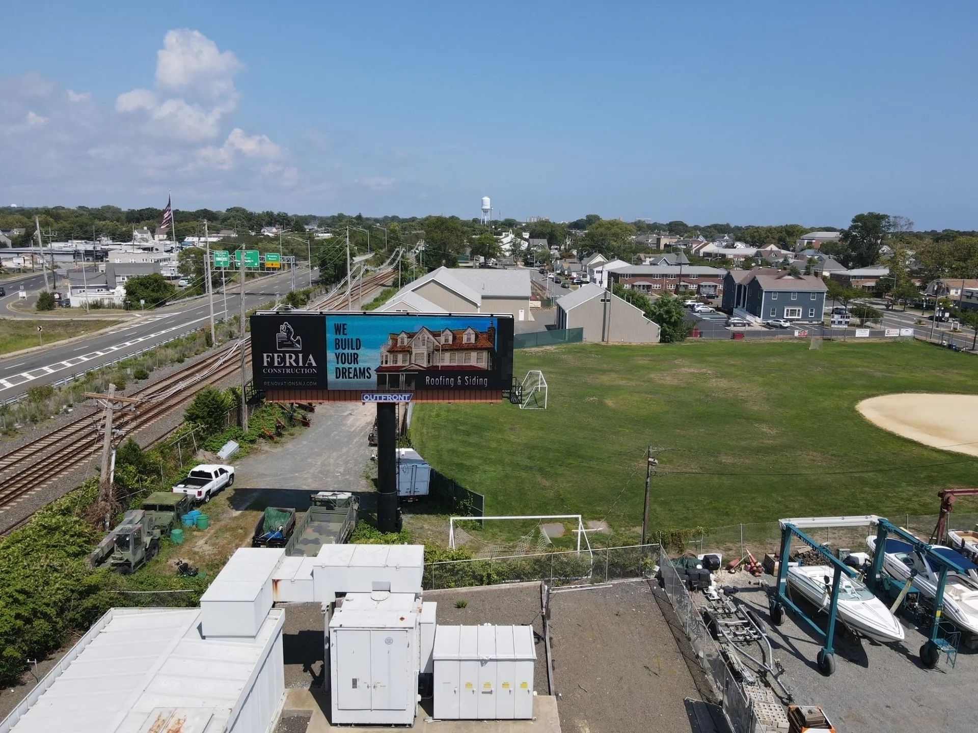 Billboard beside train tracks and a grassy lot under a cloudy sky, with utility boxes and equipment in front.