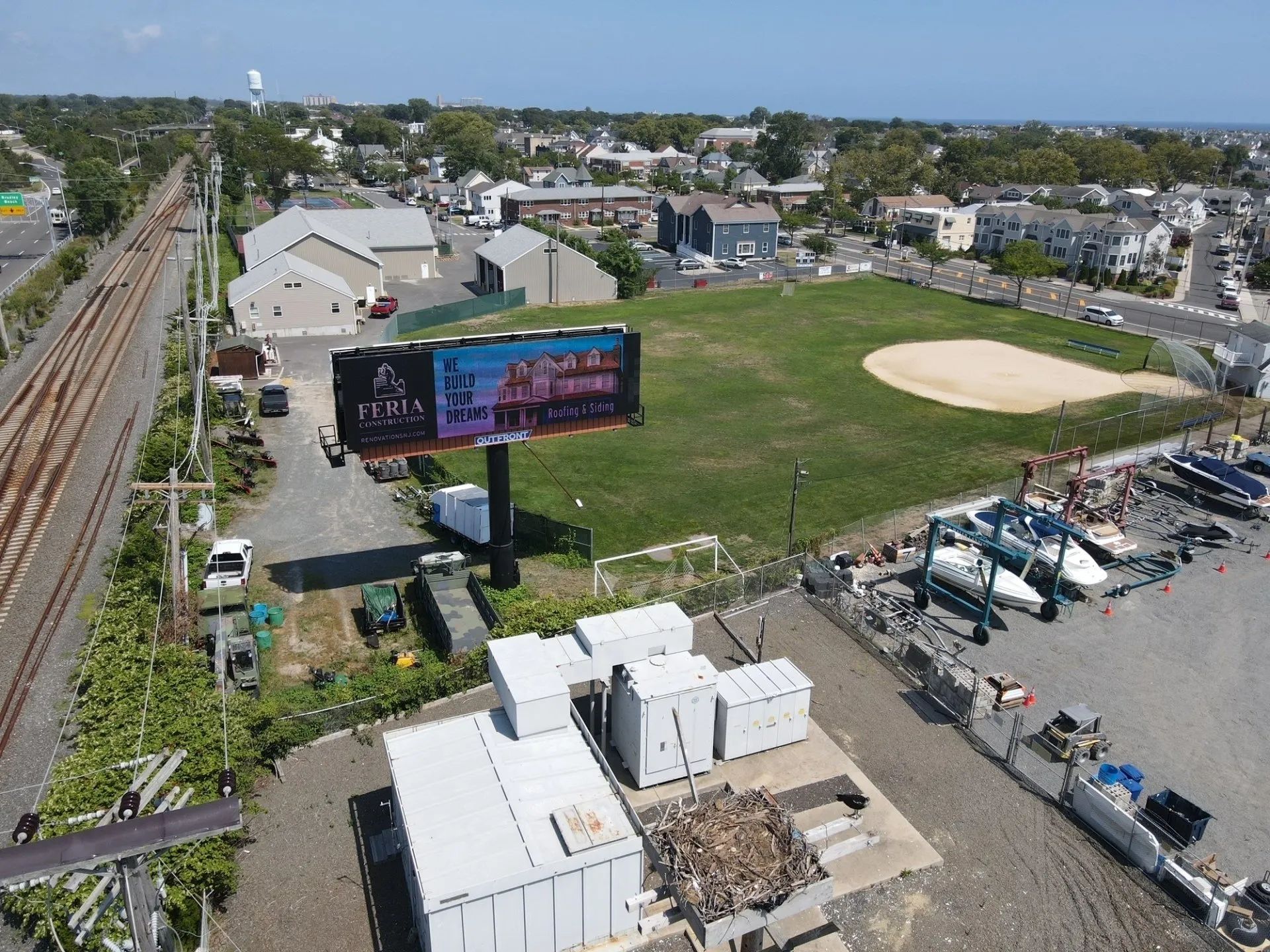 Aerial view of a baseball field beside buildings, parking lots, and railroad tracks on a sunny day