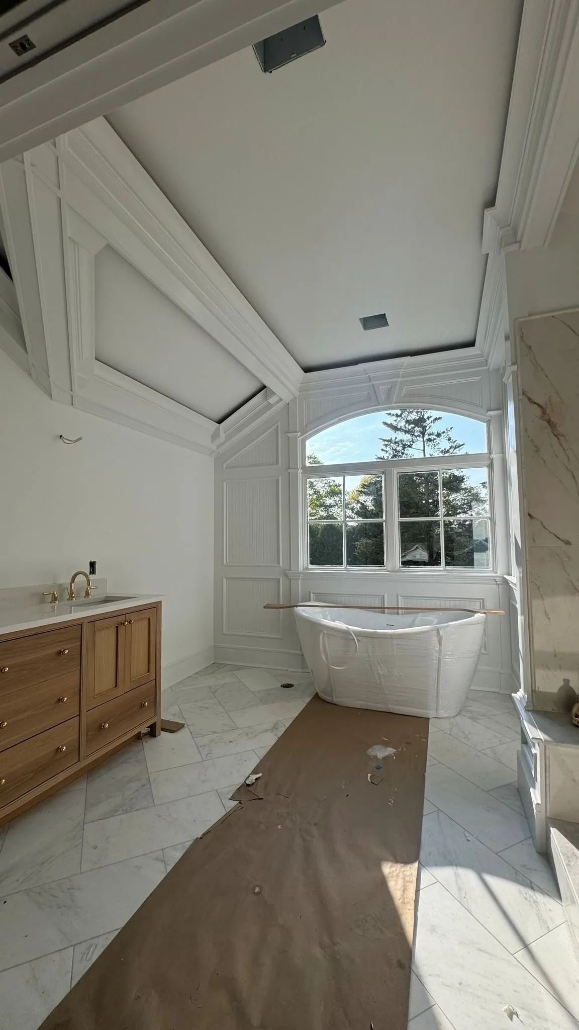 Bright white bathroom with freestanding tub, tall ceiling, wood vanity, and large window.