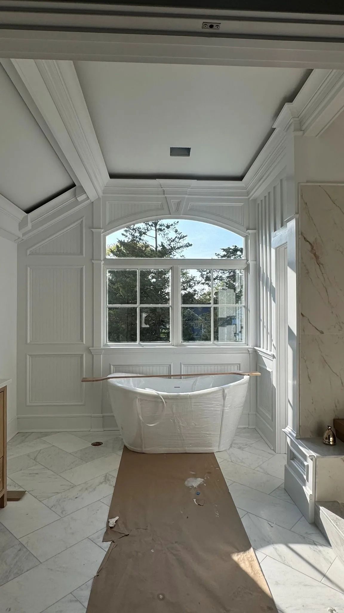 Bright white bathroom with a freestanding tub centered before a large window and ornate ceiling molding.