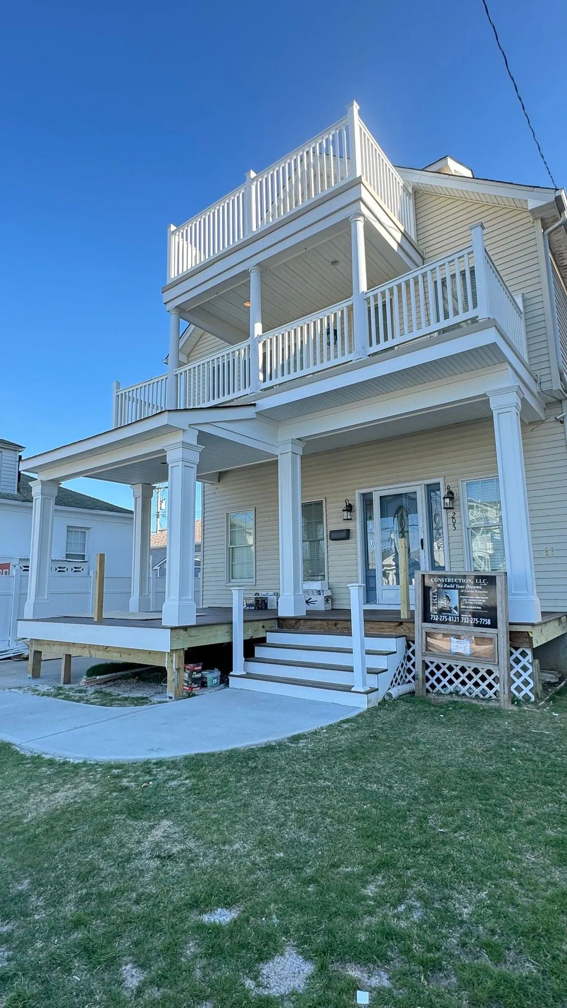 White beach house with elevated porch, balconies, and stairs, viewed from the lawn under a clear blue sky