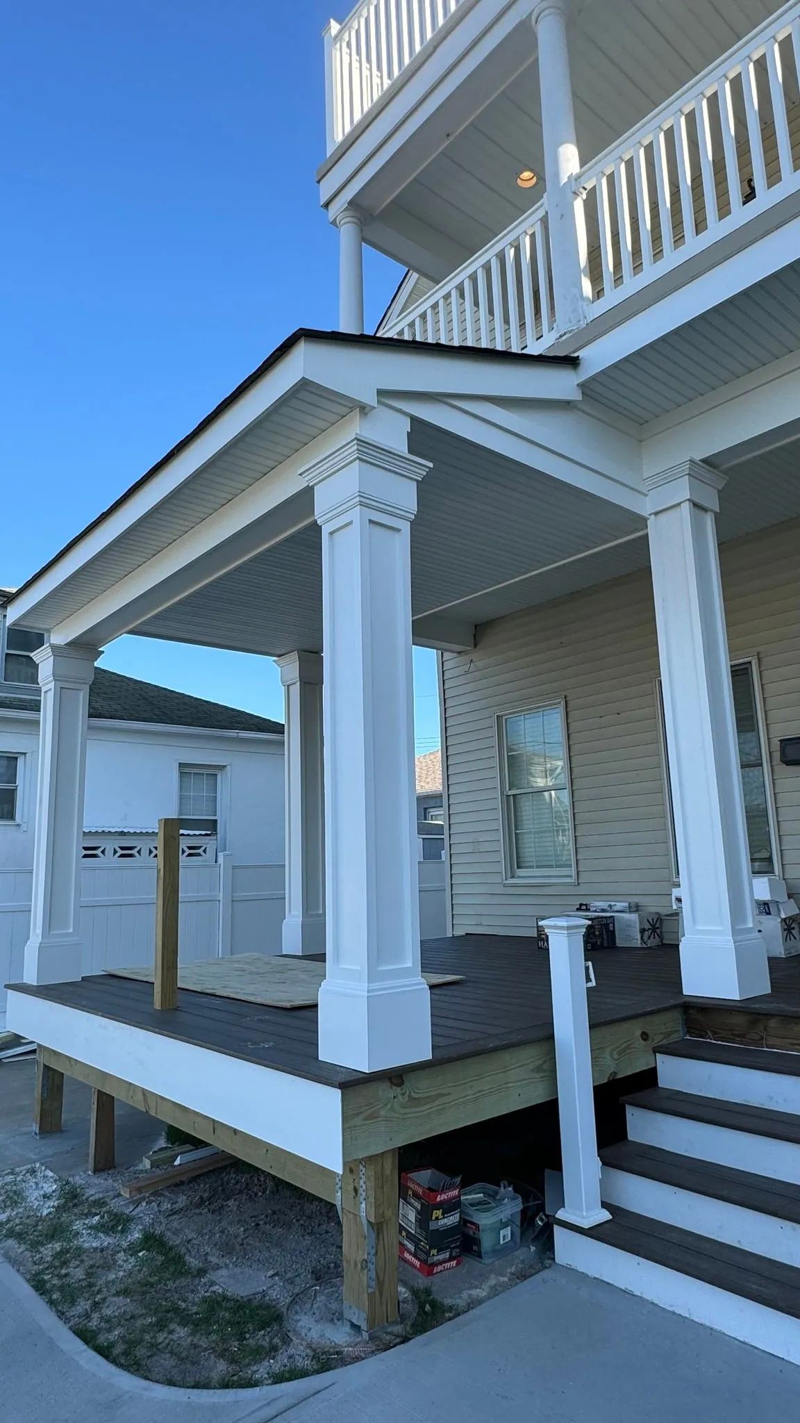 Raised house porch with white railings, stairs, and support posts beside a driveway under a blue sky