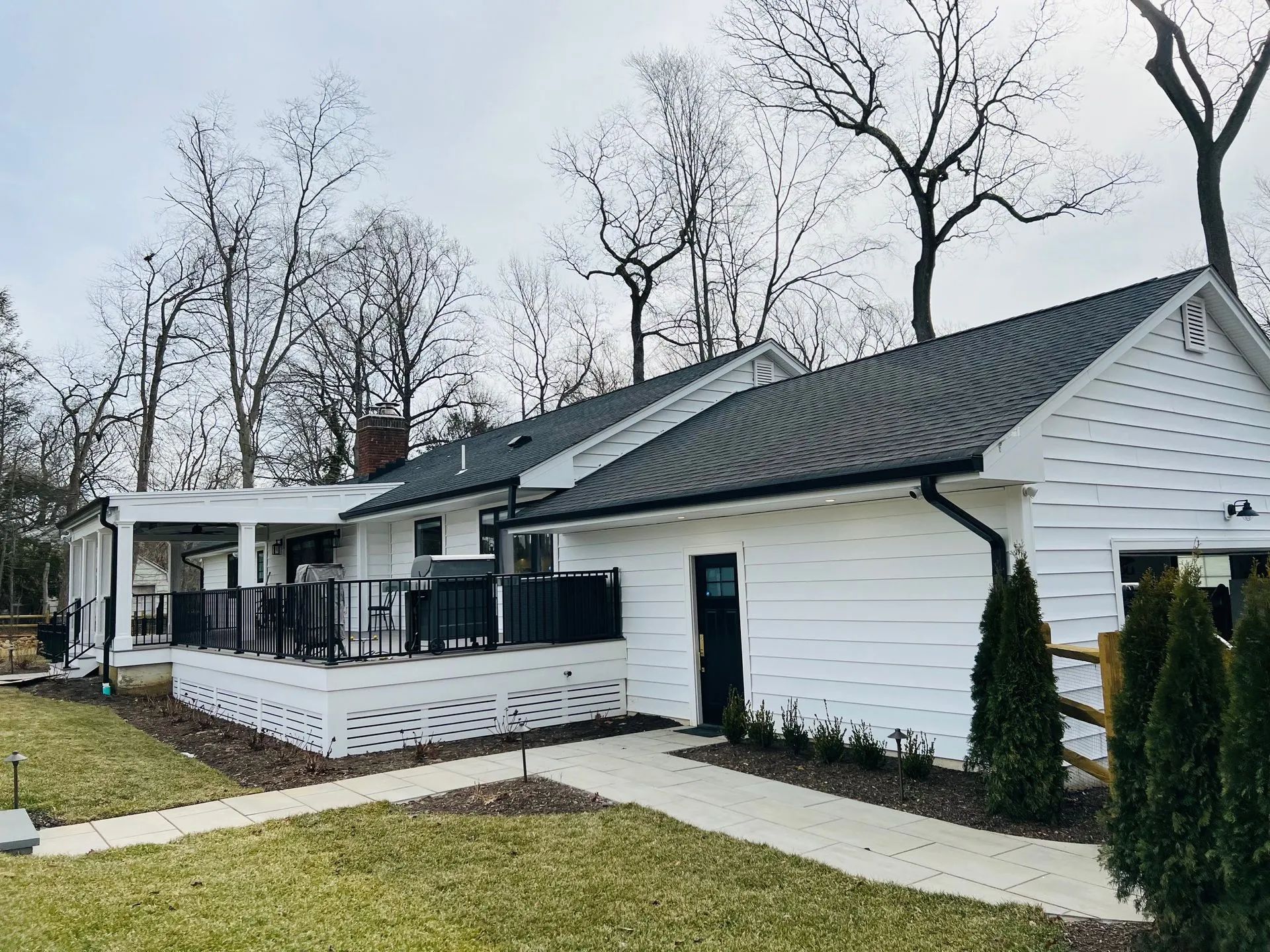 White house with dark roof, front deck, and a paved walkway in a yard with bare trees.