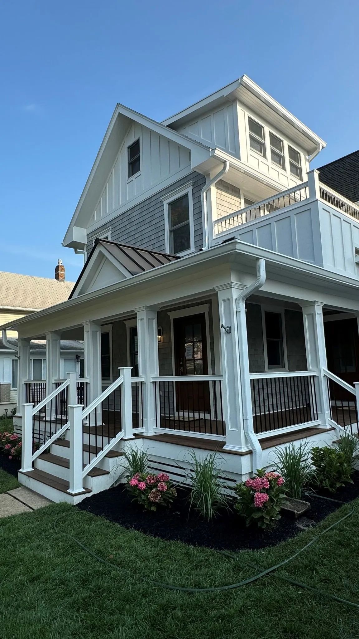Victorian-style white house with wraparound porch and flower beds under a clear blue sky