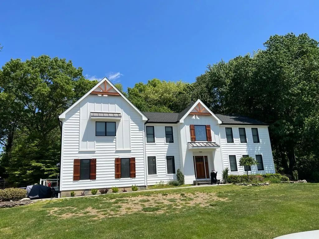 White two-story house with red shutters on a grassy hill, framed by trees under a blue sky