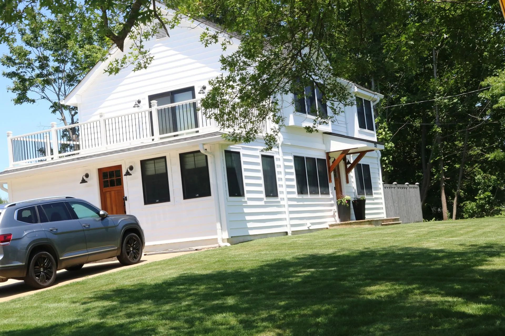 White two-story house with a balcony, a silver SUV in front, and trees shading a grassy yard