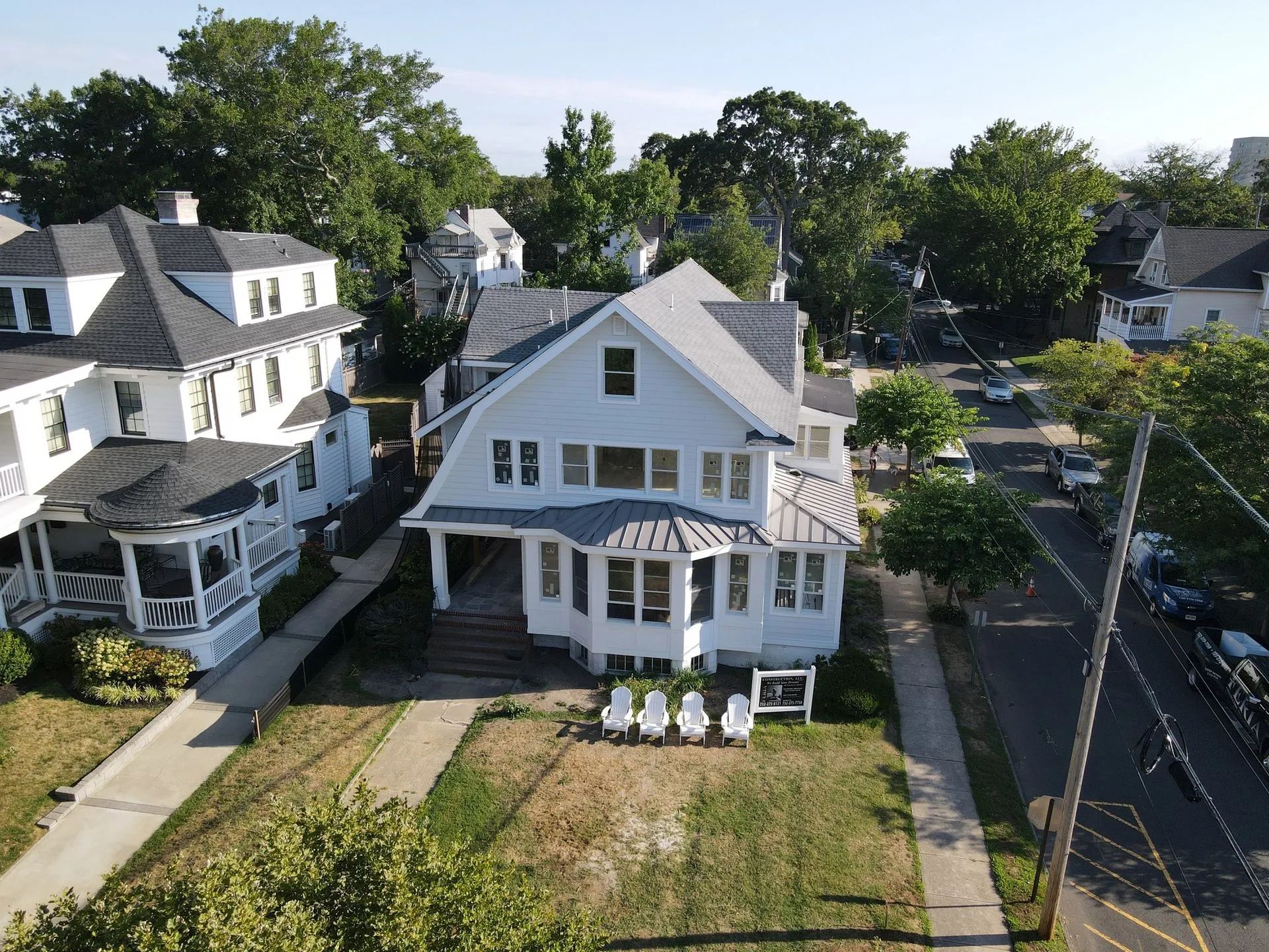 Aerial view of a white house with a porch, front yard, and neighboring homes along a tree-lined street.