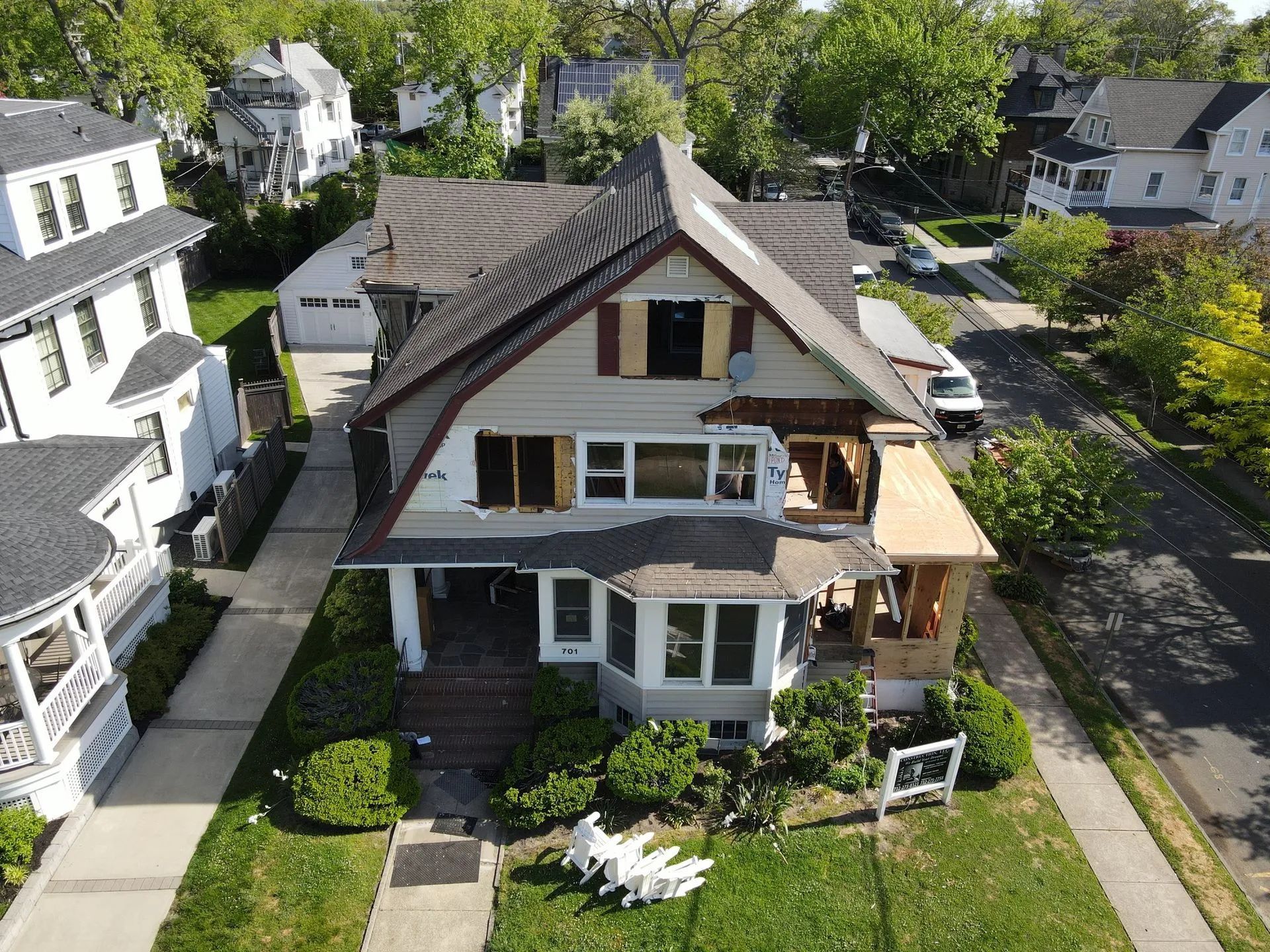 Aerial view of a two-story house with a front porch, brown roof, and a small landscaped yard on a sunny street.