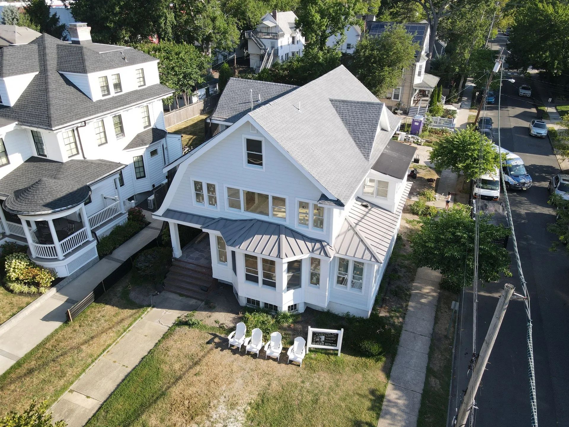 Aerial view of a white house with a porch, lawn chairs, and a driveway beside a tree-lined street.