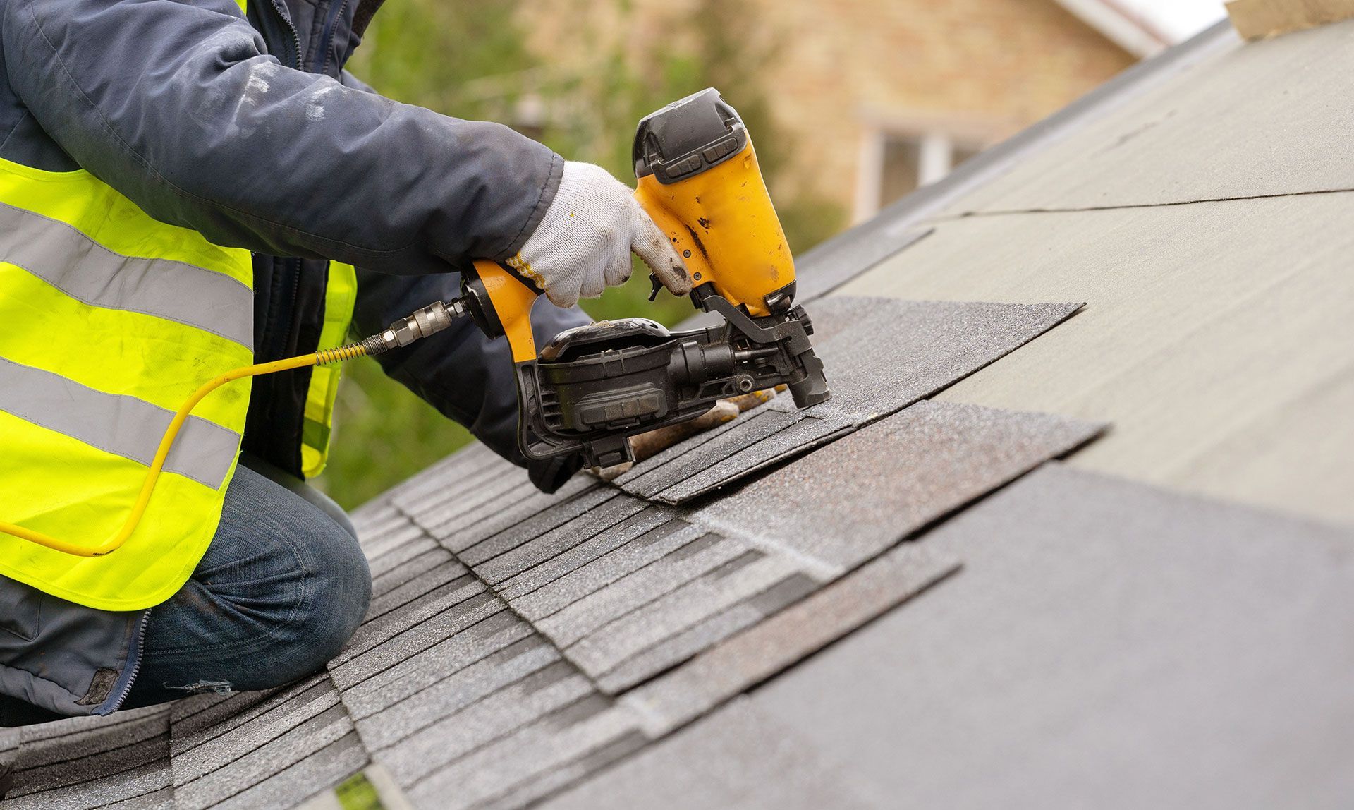 Worker in a yellow safety vest using a nail gun to install roof shingles on a sloped roof