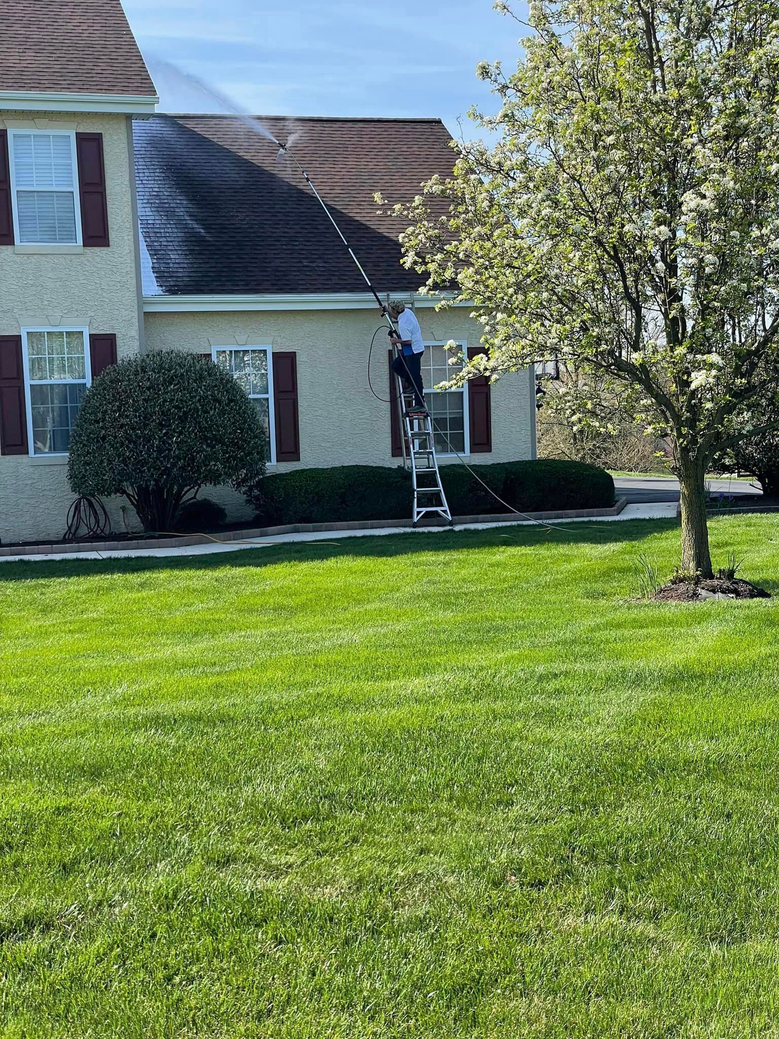 A man is standing on a ladder cleaning the roof of a house.