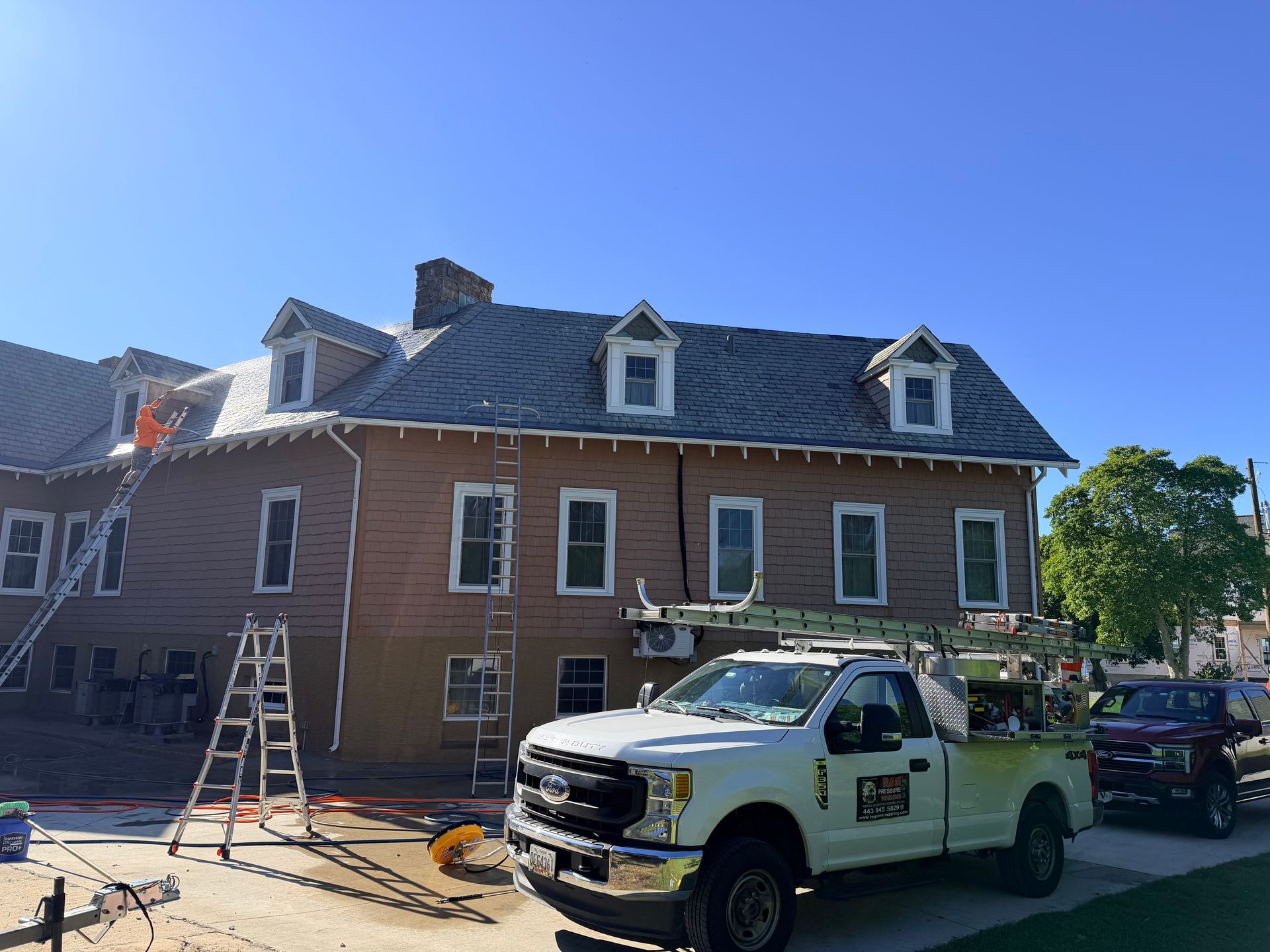 A white work truck parked in front of a building with ladders leaning against it; roofers are visible on the roof.