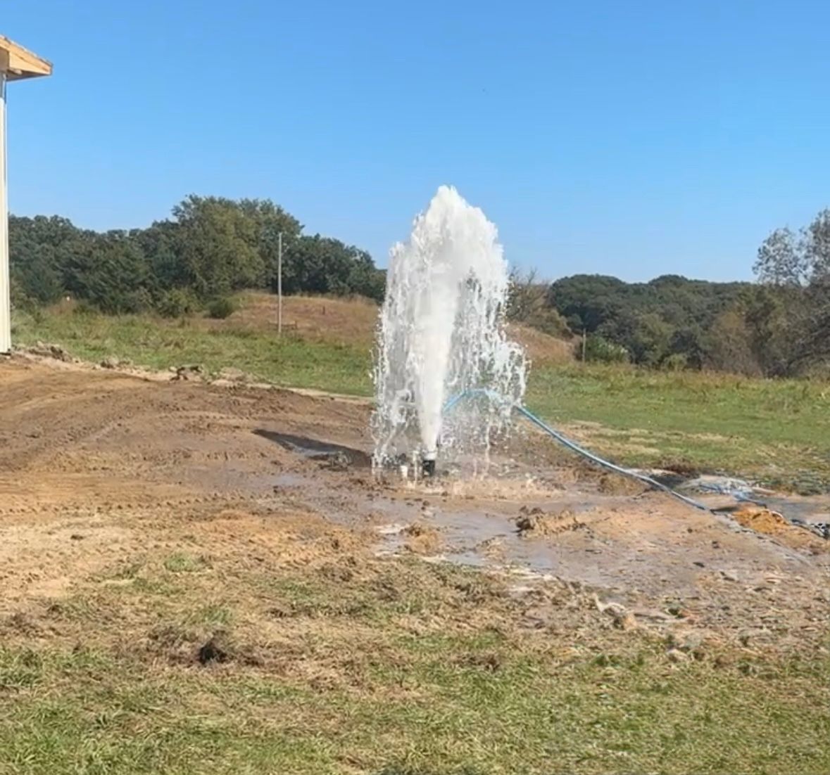 A geyser of water erupts from a well in a grassy field under a clear blue sky.