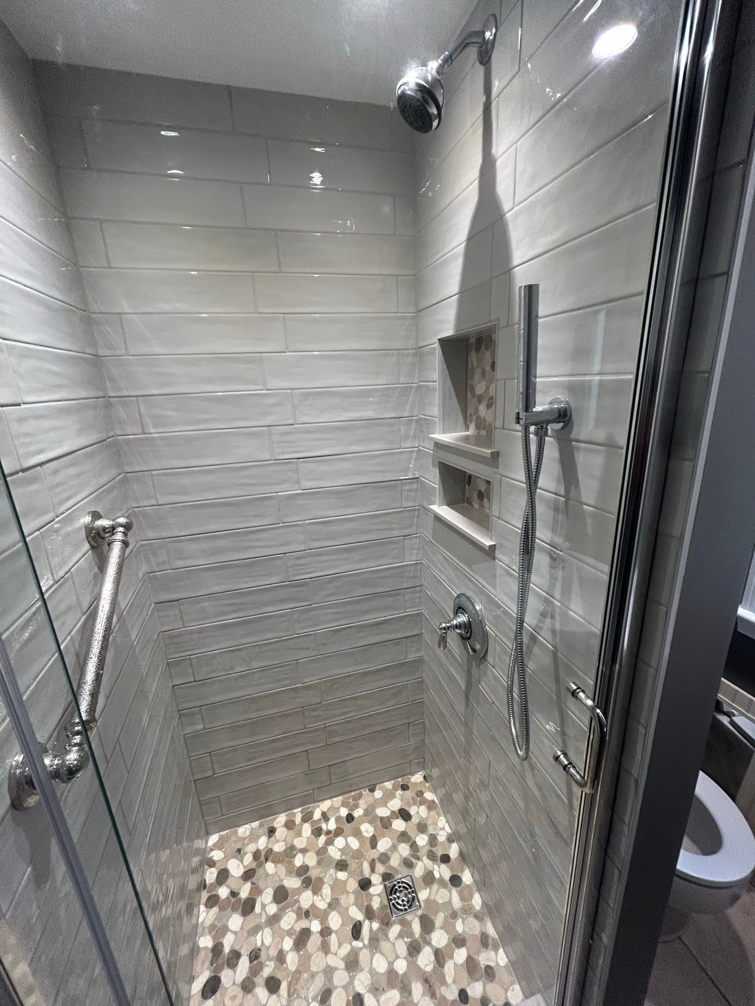Small, modern shower with gray tile walls, pebble floor, and glass door.
