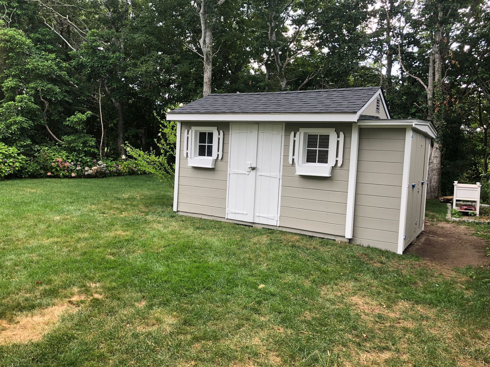 Tan shed with white doors, window shutters, and a dark roof, in a grassy backyard, surrounded by trees.