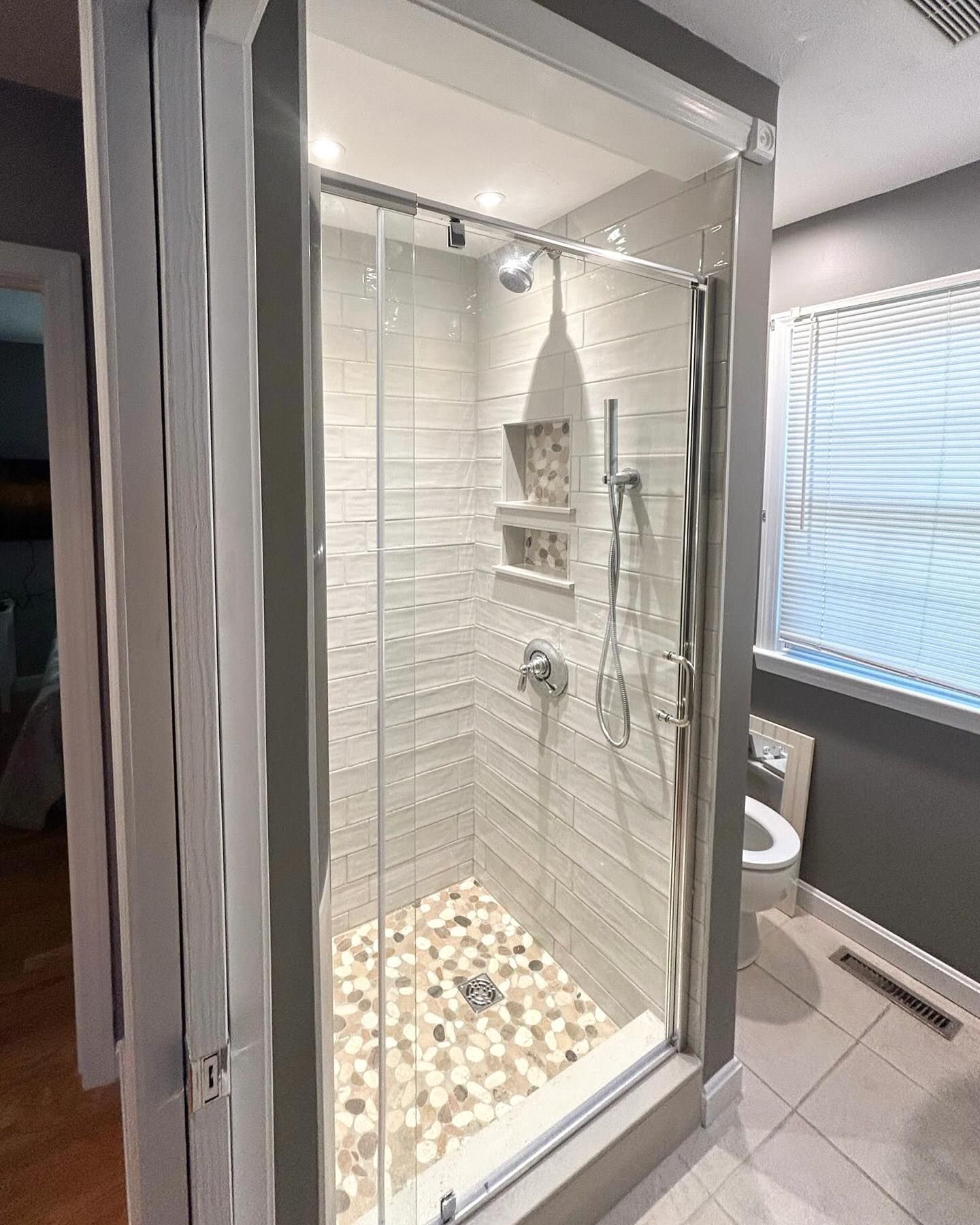 Modern shower with glass door, white textured tile, pebble floor, and built-in shelving.