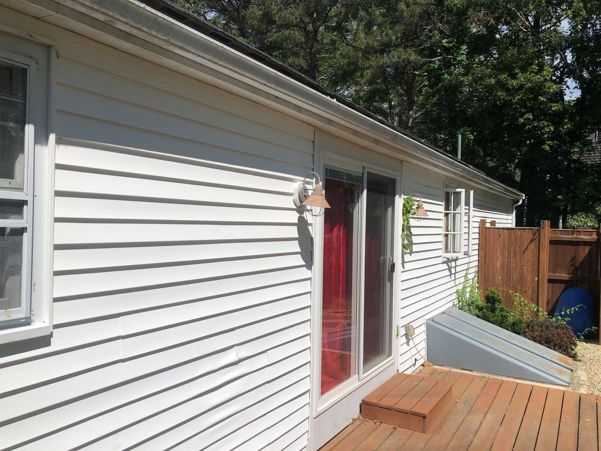 White-sided house with a red door, wooden deck, and a window. Sunny outdoor setting.