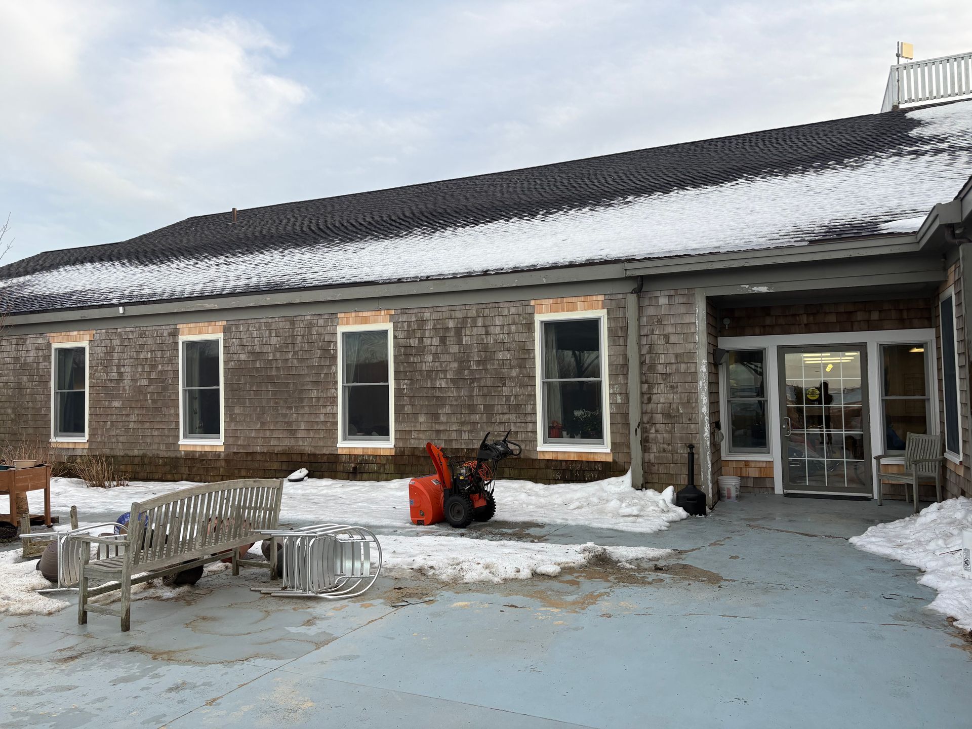 A single-story stone building with a snow-covered roof and patio, featuring a bench and a snow blower by the entrance.