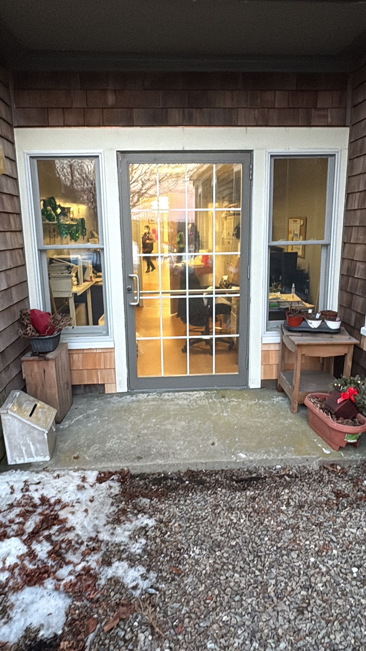 A building entrance with a glass door, flanked by two windows and decorated with small tables and a plant.