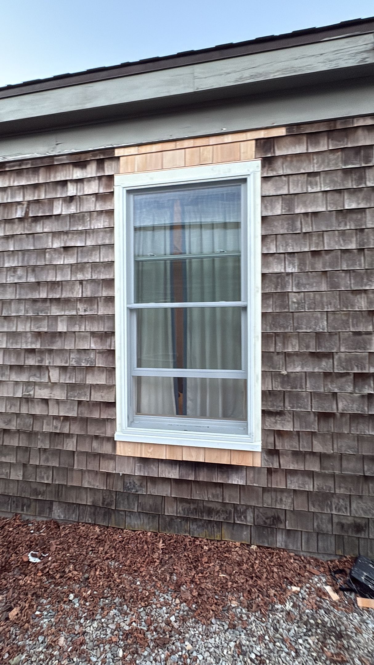 A newly installed white-framed window sits in a wall of weathered cedar shingles, with fresh, light wood trim around it.