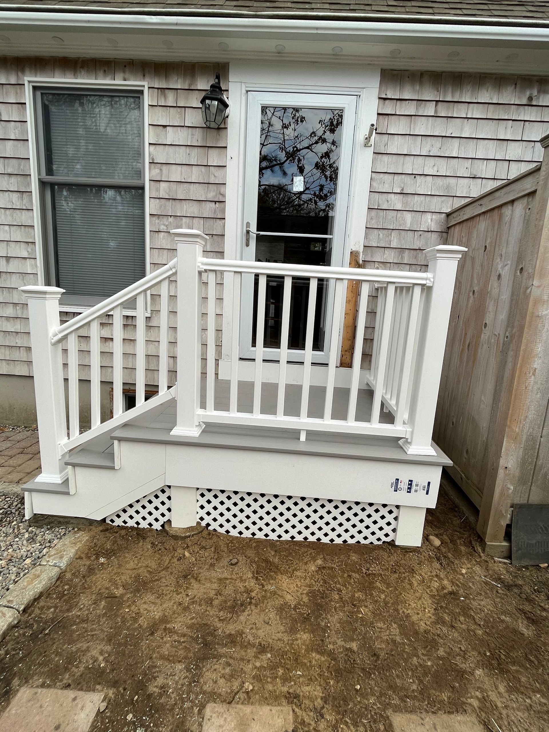 A wooden porch with a white railing and lattice skirting, accessed by two small steps leading to a glass-paneled door.