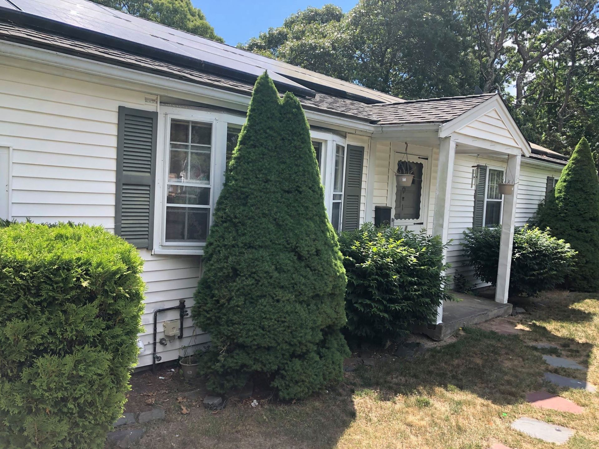 White house with dark roof and solar panels, green shutters, bushes, and a pathway.